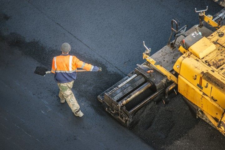 A worker in a high-visibility orange jacket uses a shovel to smooth fresh black asphalt behind a paving machine.