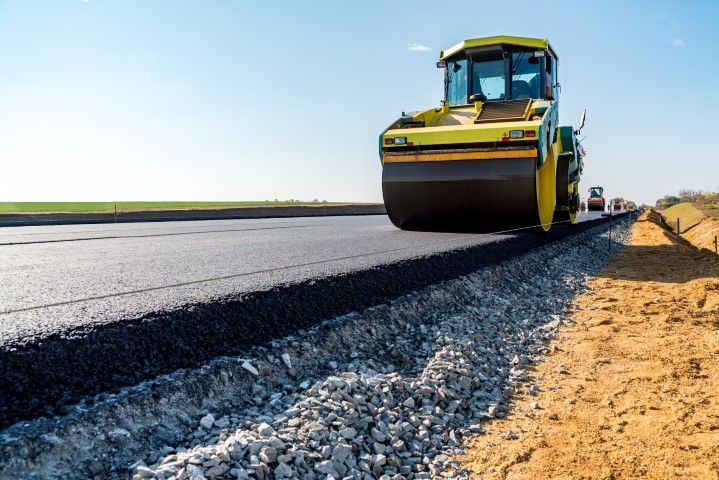 A yellow construction road roller compacts freshly laid dark asphalt on a highway project beside a gravel shoulder.