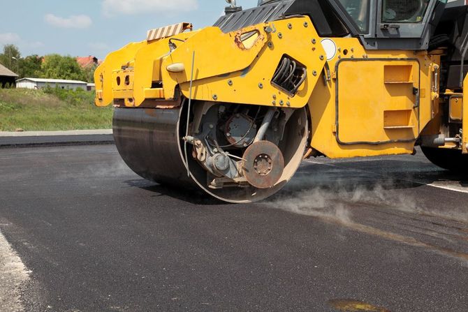 A yellow steamroller compacts fresh, dark asphalt on a road construction site.