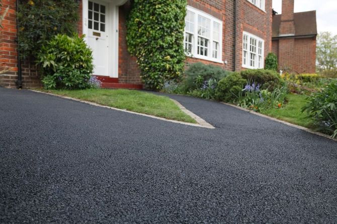 A freshly paved black asphalt driveway leads to the white front door of a brick house with manicured green landscaping.