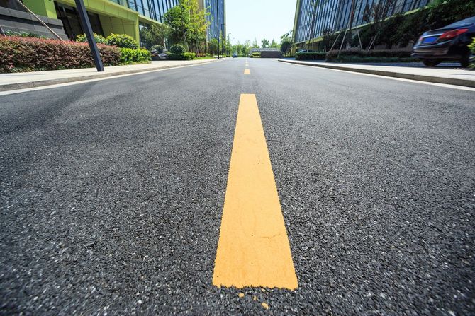 A low-angle view of an asphalt road with a bright yellow centerline, flanked by green bushes and modern office buildings.