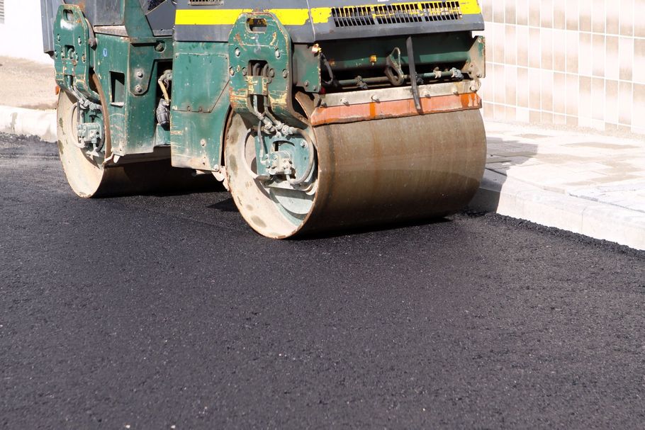 A green road roller flattens a fresh layer of black asphalt beside a concrete curb.