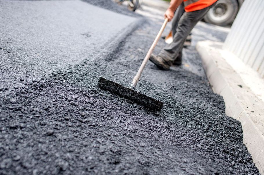 A construction worker in an orange vest spreads fresh asphalt on a road surface using a long-handled rake.