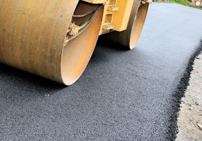 A large yellow industrial road roller compacting a layer of freshly laid dark asphalt on a road.