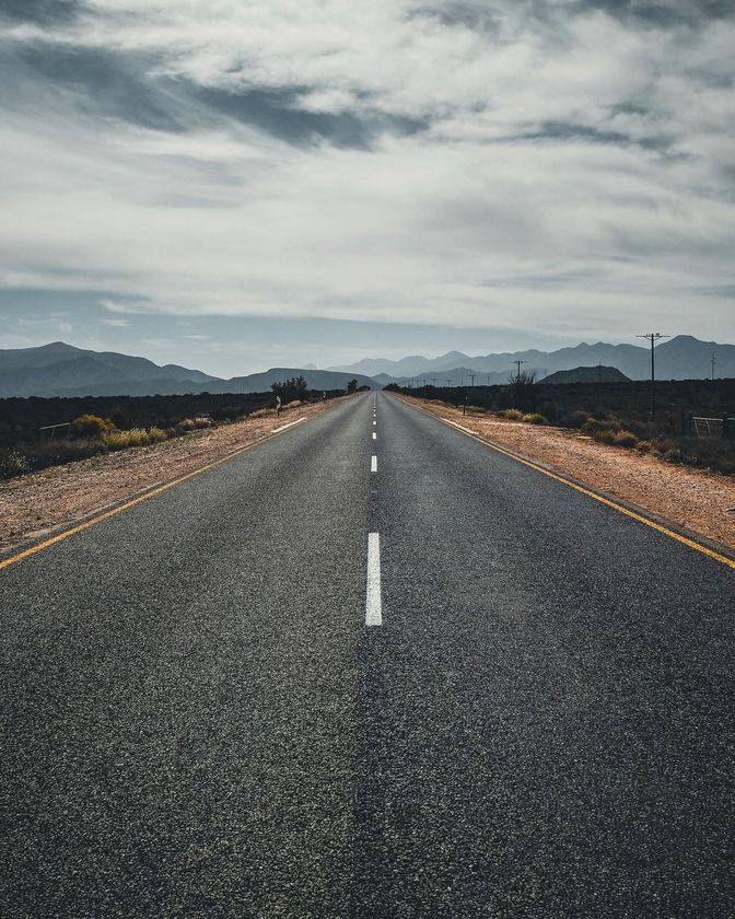 A long, empty asphalt road stretches toward distant mountains under a cloudy, overcast sky.