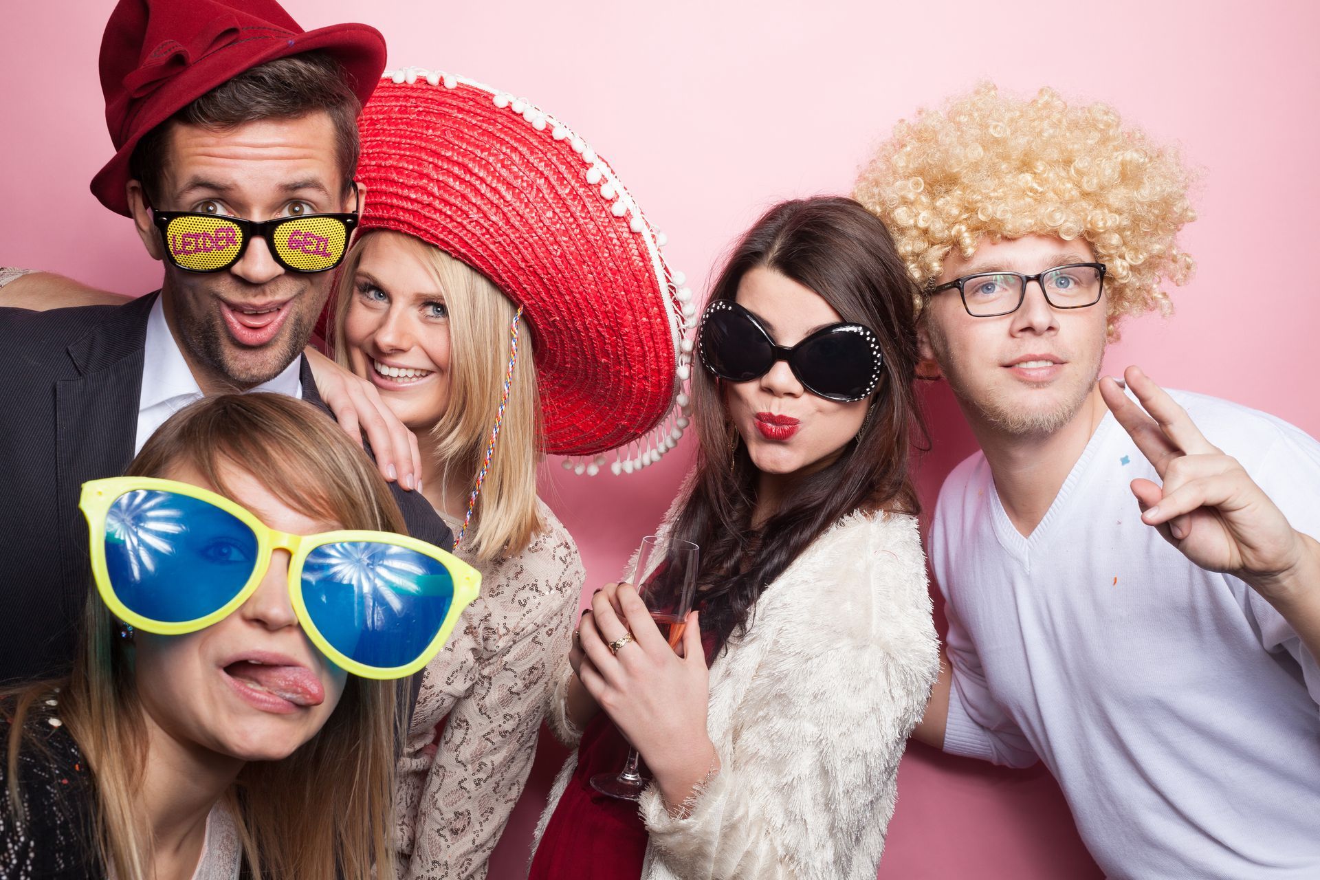 Four people wear festive props, including hats, wigs, and oversized sunglasses, while posing against a pink background.