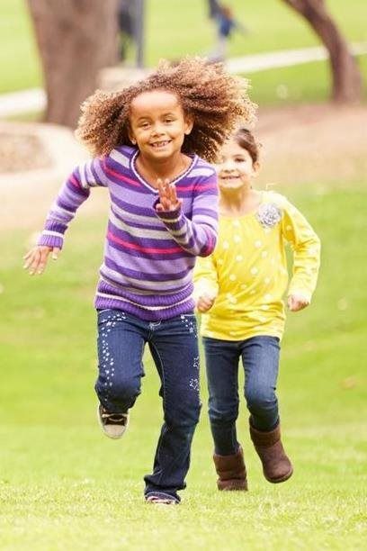 Two children running on grass, smiling, with trees in the background.