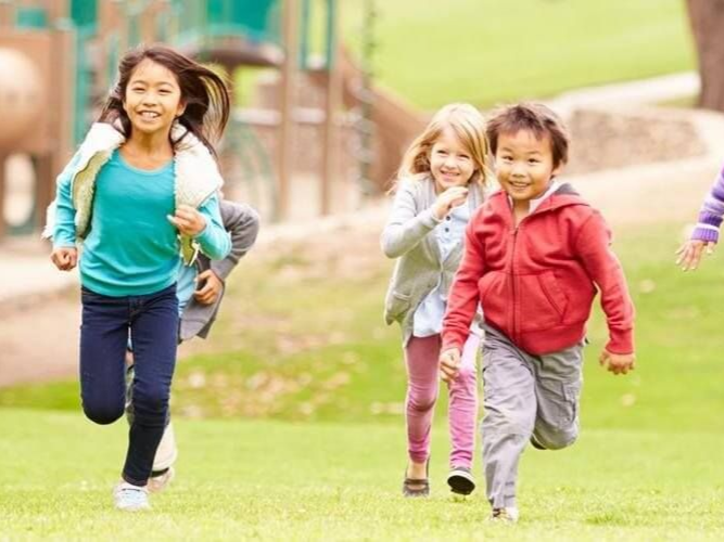 Children running and smiling on a grassy field, one in a red jacket and one in turquoise.
