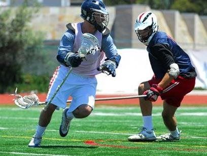 Two lacrosse players in blue and black gear compete on a turf field, one cradling the ball.