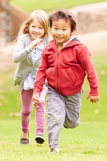 Two children running on grass outdoors, smiling, one in a red hoodie and one in a gray sweater.