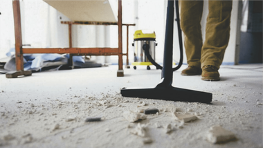 Construction dust on a floor being swept with a broom; a person’s legs stand nearby.