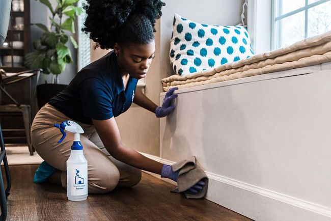 Person kneeling and cleaning a white baseboard with a spray bottle in a living room