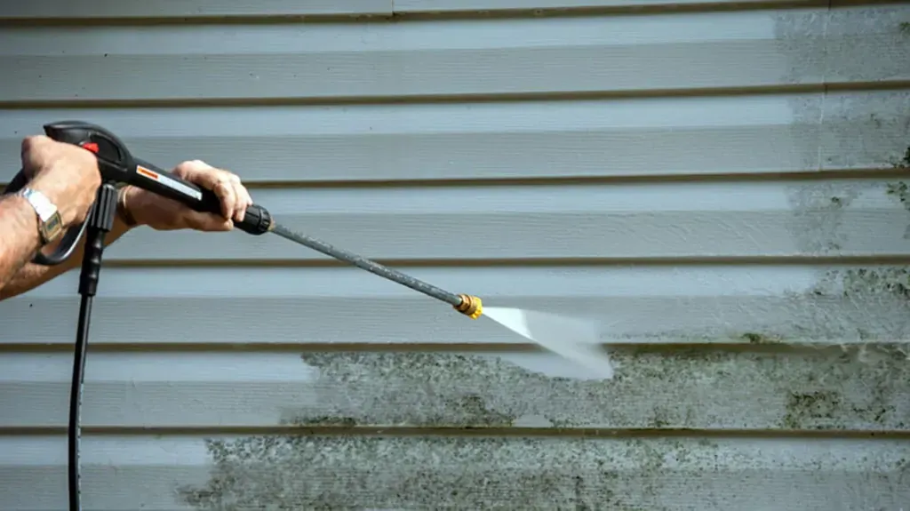 Person pressure-washing a gray vinyl wall with a spray wand