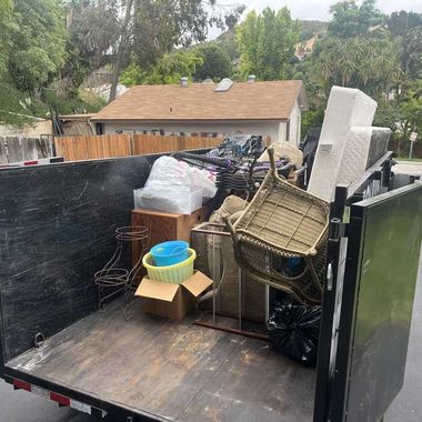 Dumpster filled with mixed household items and furniture, including a wicker chair and boxes, in a driveway.