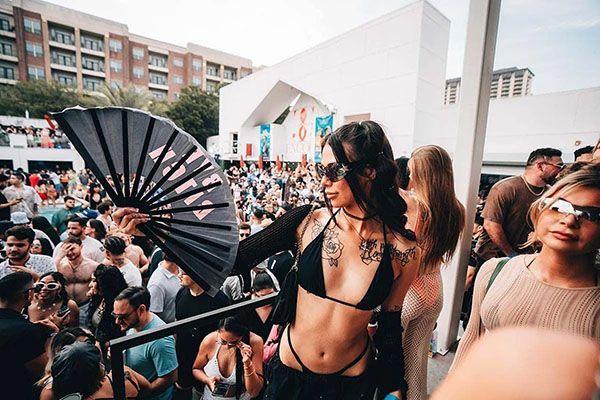 Woman at a crowded outdoor event, holding a fan. Tattoos, sunglasses, and a black bikini top.