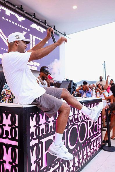 A performer in a white t-shirt and grey shorts sits on a stage edge, gesturing with a microphone during an event.