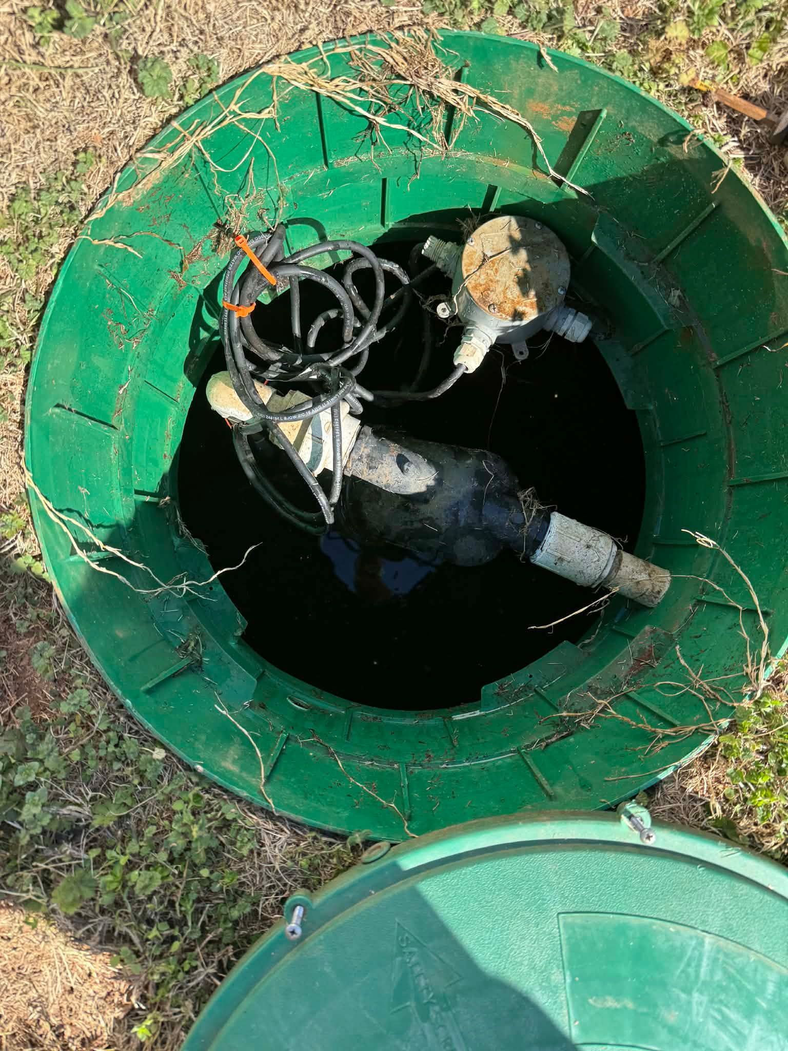 Top-down view of an open green septic pump basin, showing submersible pumps, pipes, and electrical cables inside.