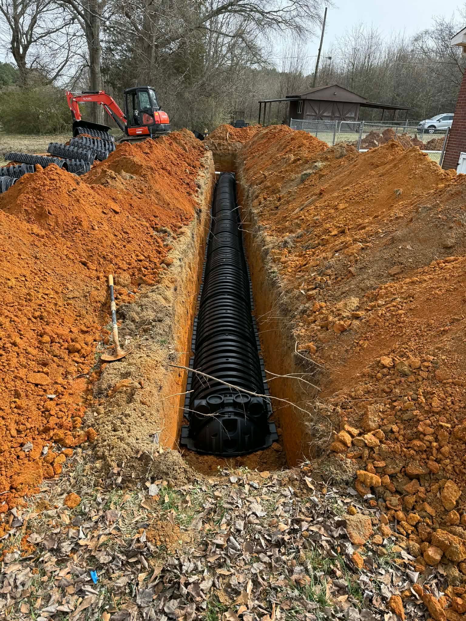 A black corrugated plastic septic chamber installed in a deep trench lined with reddish-brown soil, with an excavator nearby.