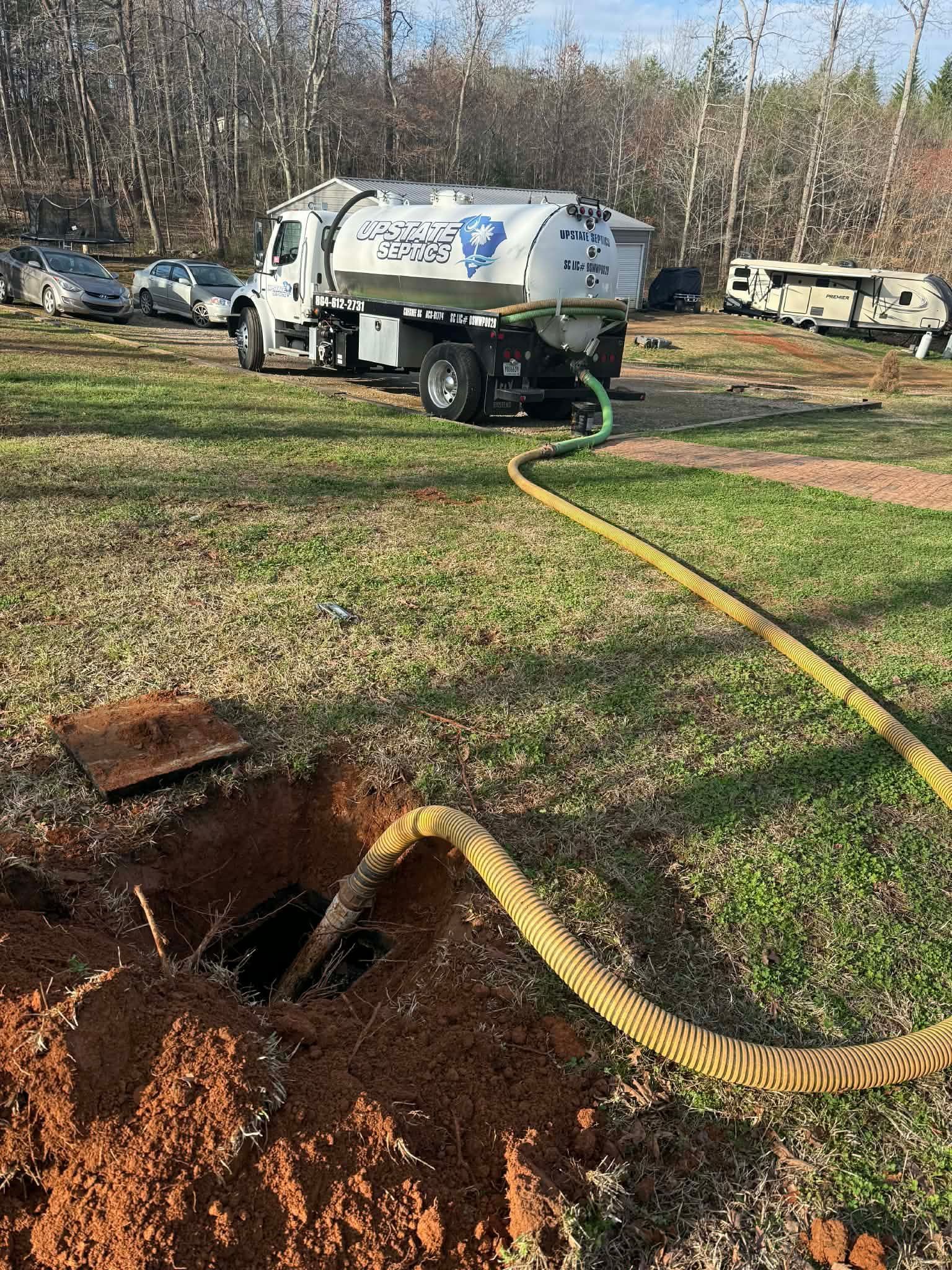 A white septic service truck is parked on grass, pumping out an underground tank via a long, yellow corrugated hose.