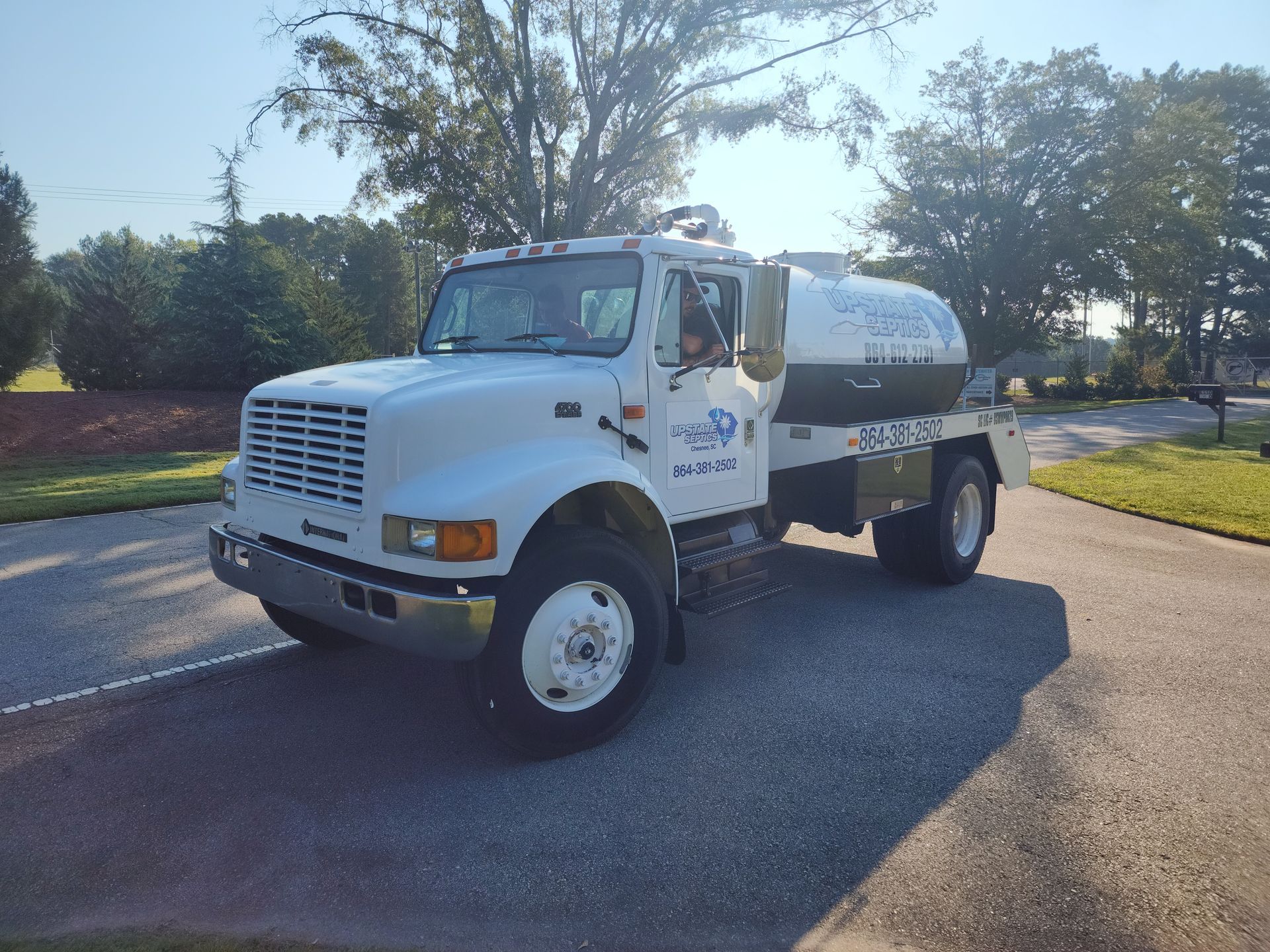 A white, commercial tanker truck parked on an asphalt surface during a sunny day with trees in the background.
