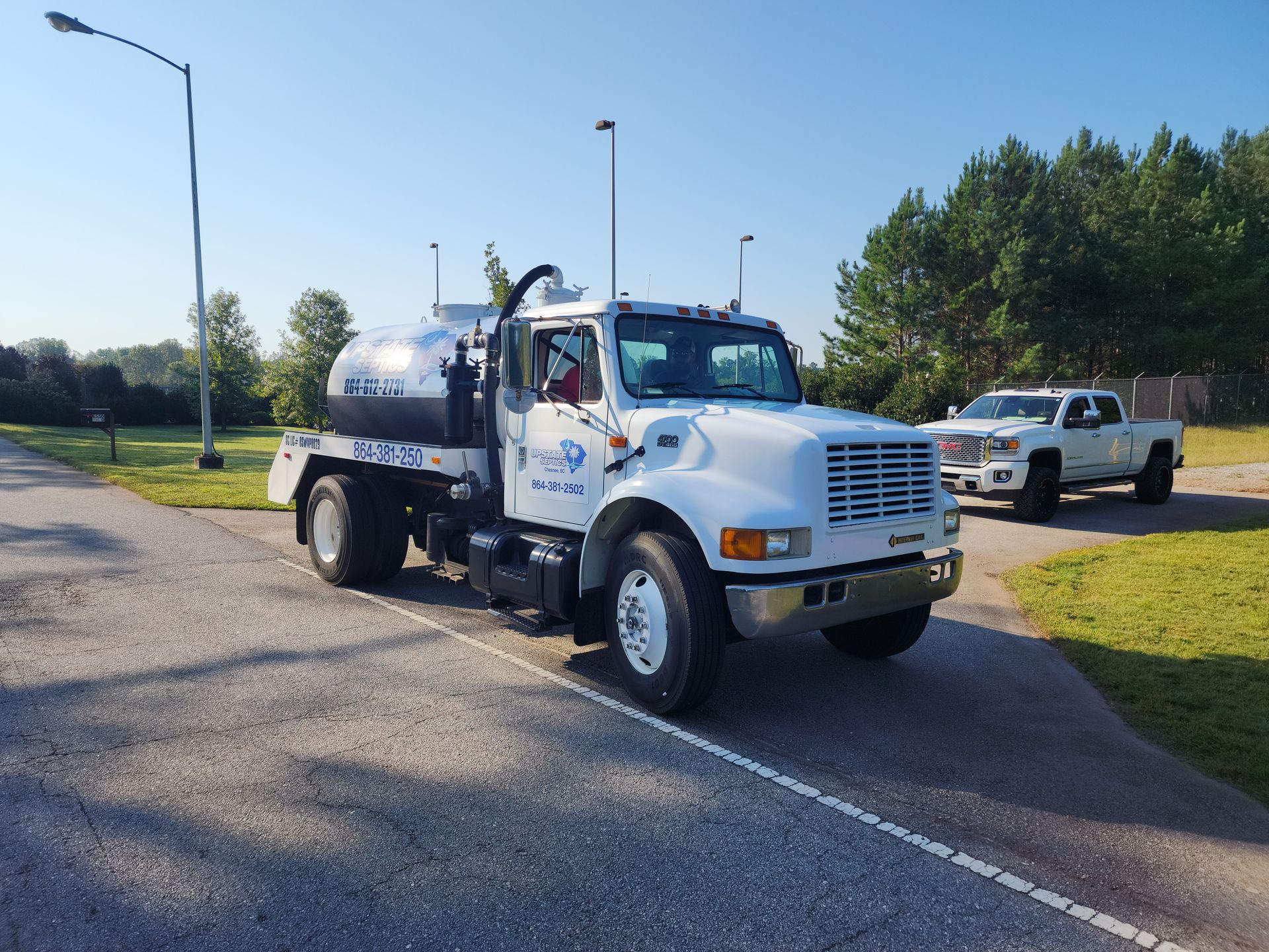 A white utility truck with a large tank and a matching pickup truck parked on an asphalt lot near trees.
