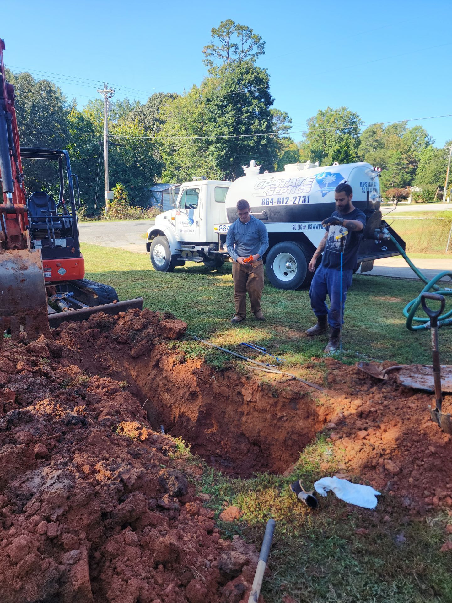 Two workers stand by a deep trench and an excavator next to a service truck on a sunny day.