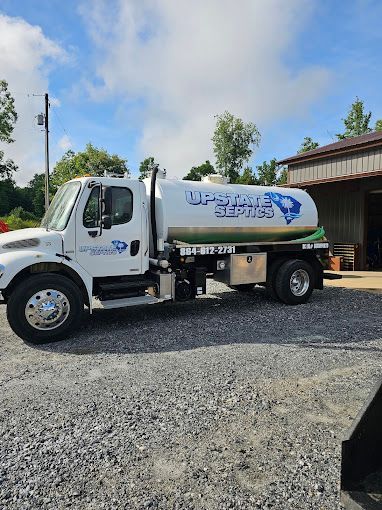 A white Upstate Septic service truck parked on a gravel lot near a metal building under a sunny blue sky.