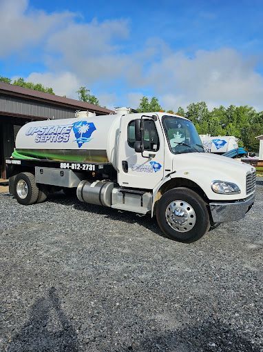 A white Up-State Septic tanker truck parked on a gravel lot under a sunny blue sky.