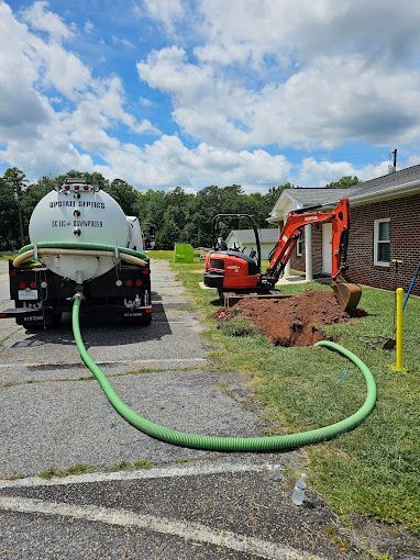 A vacuum truck with a green hose connected to an excavated hole next to an orange excavator in front of a brick building.