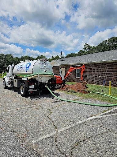 A vacuum truck pumps a trench outside a brick building while a small orange excavator is parked nearby.