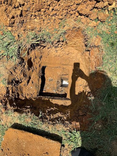 A hole in the ground reveals a square, dark container with a light-colored pipe fitting inside.