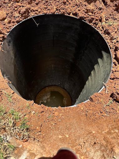 A deep, black circular pipe or shaft is partially buried in reddish-brown dirt, with a small pool of water at the bottom.