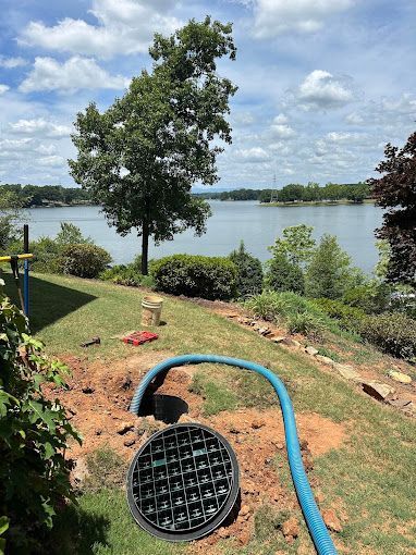 A black drainage grate installed in a grassy yard overlooking a lake, with a blue hose connected to it.
