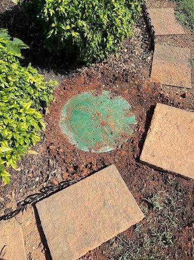 A green, circular irrigation valve box cover sits in the dirt next to stone garden path pavers and a bush.