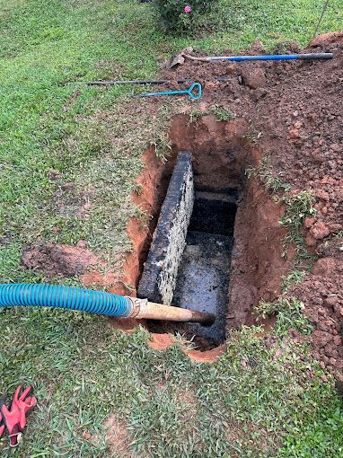 A blue suction hose extends into a deep, rectangular hole in the ground to pump out a residential septic tank.