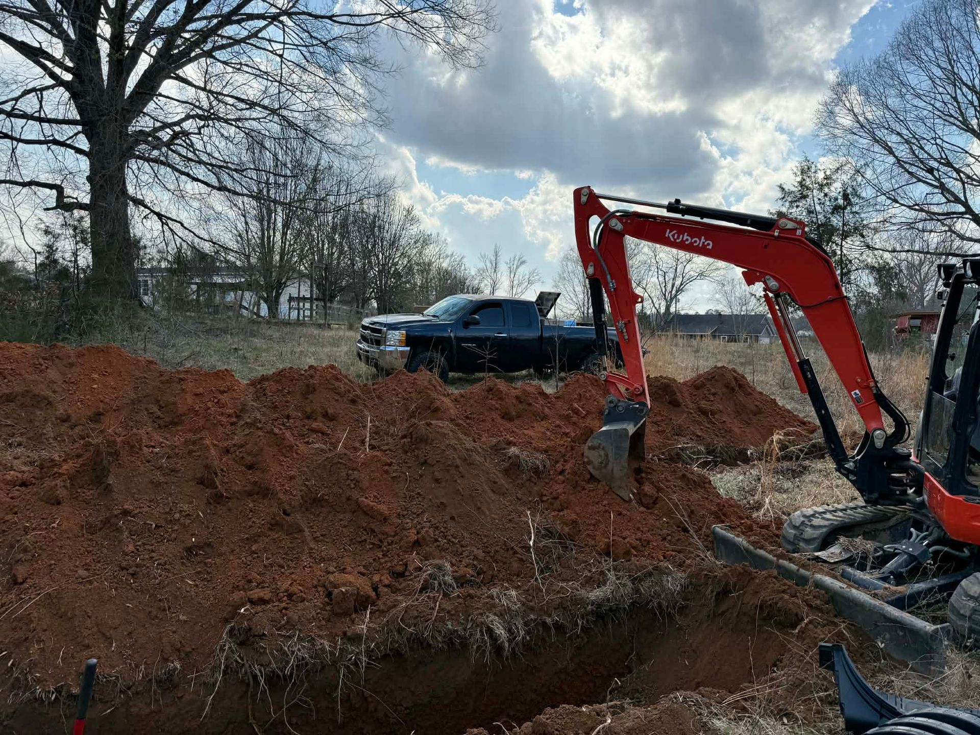 A red excavator digs a trench in a field with a dark truck parked in the background under a cloudy sky.
