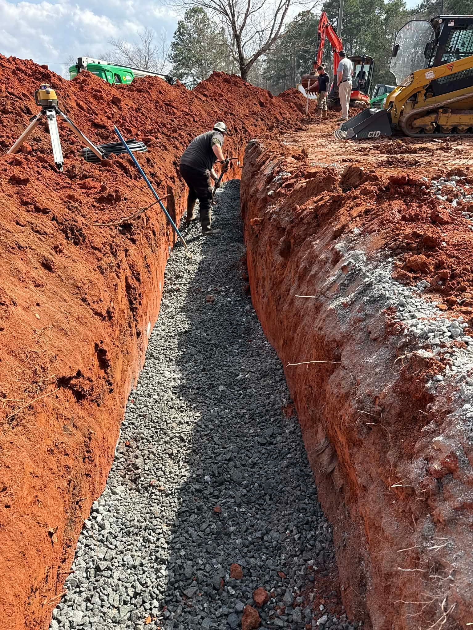 A construction worker stands in a deep trench lined with gravel, with a yellow skid steer and machinery in the background.