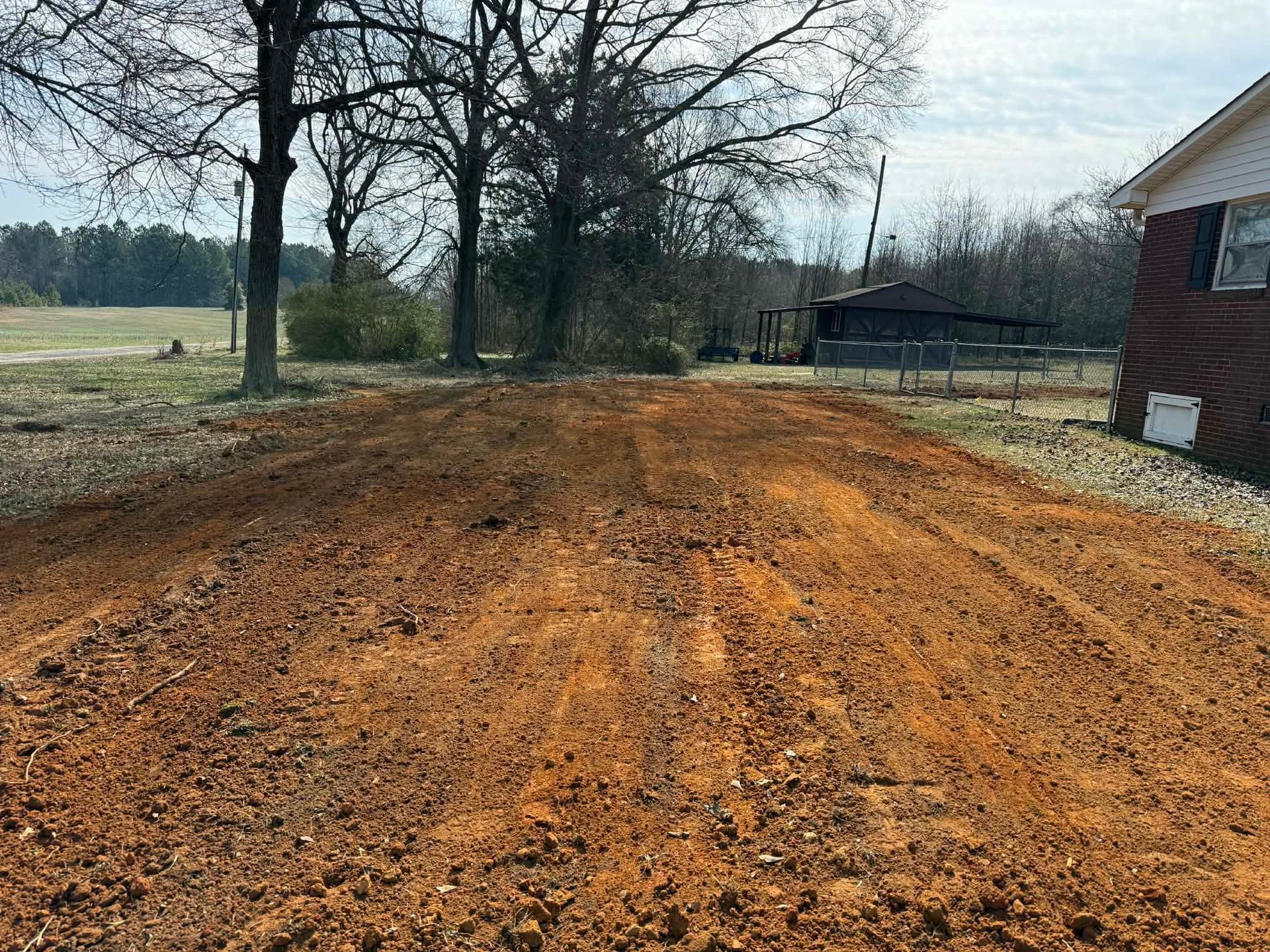 A freshly tilled, reddish-brown dirt yard extending toward trees and a house on a clear, sunny day.
