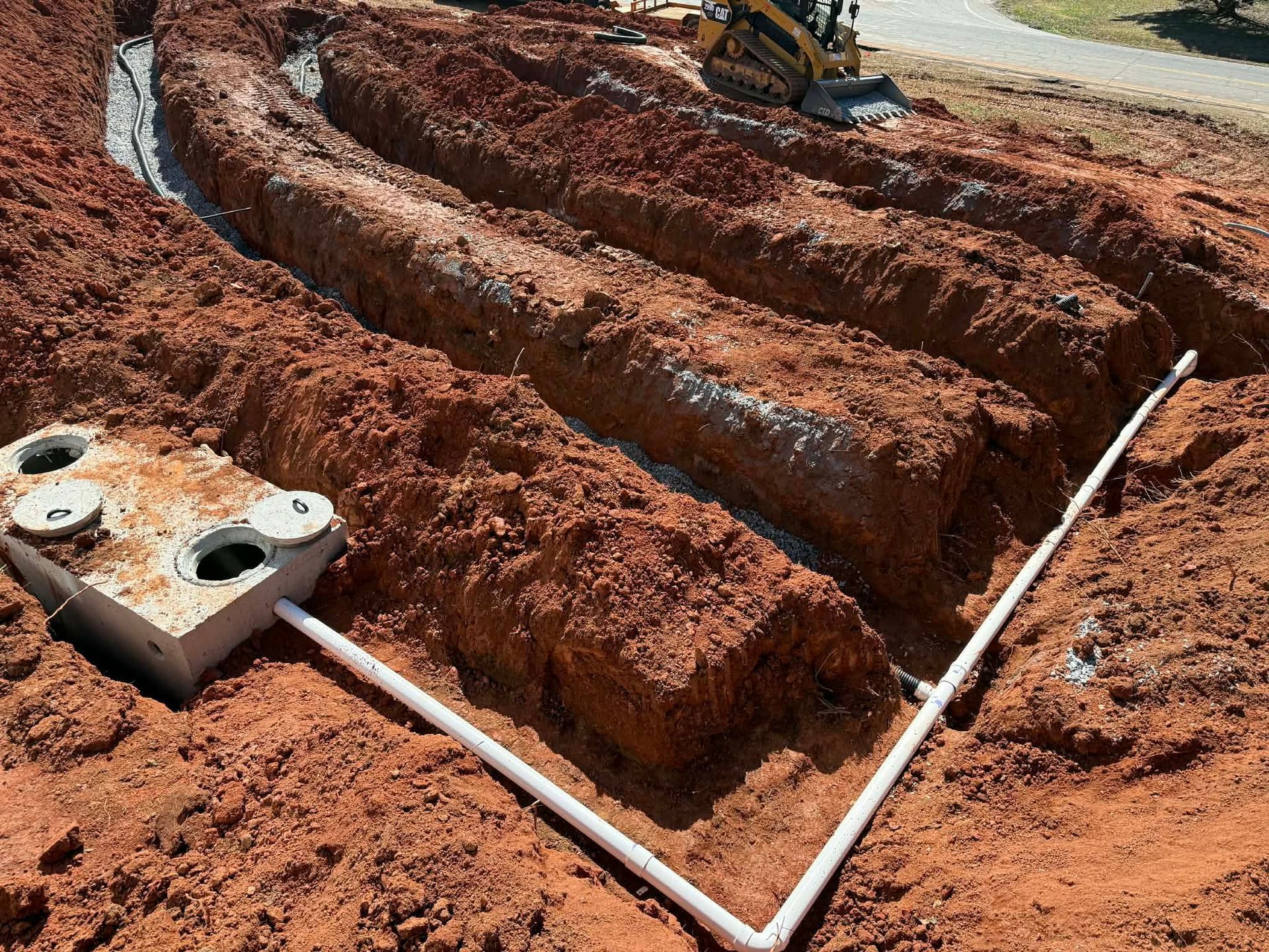 An active construction site showing a concrete septic tank connected to a series of excavated trenches in red clay soil.