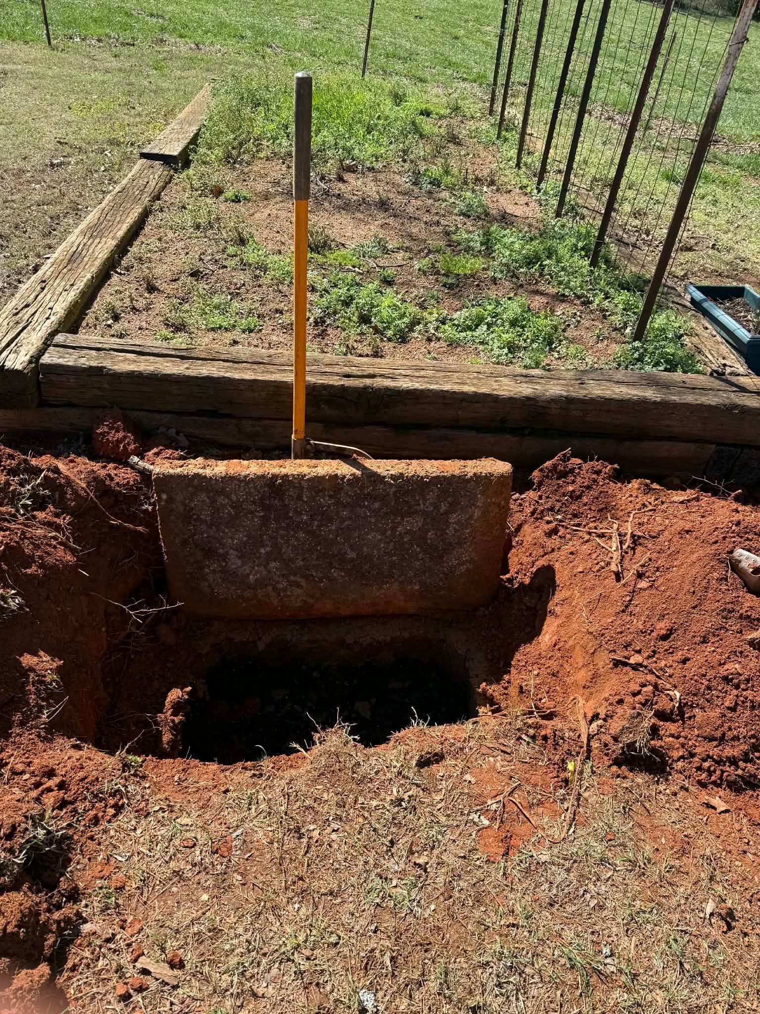 A rectangular concrete septic tank lid sits half-open in a dug-out hole of red dirt next to a raised wooden garden bed.