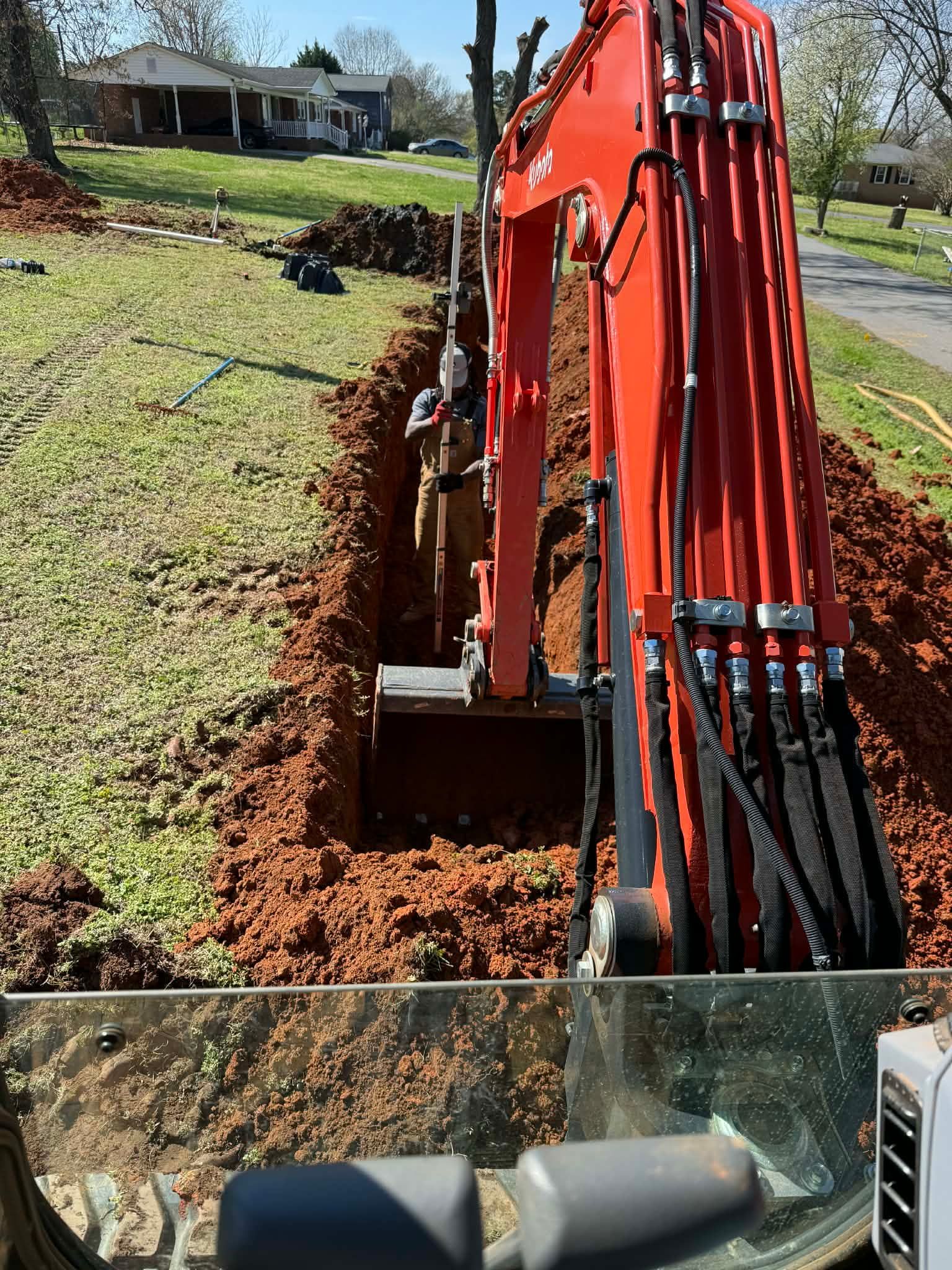 An orange excavator digging a deep trench in a yard, with a worker standing inside the trench.