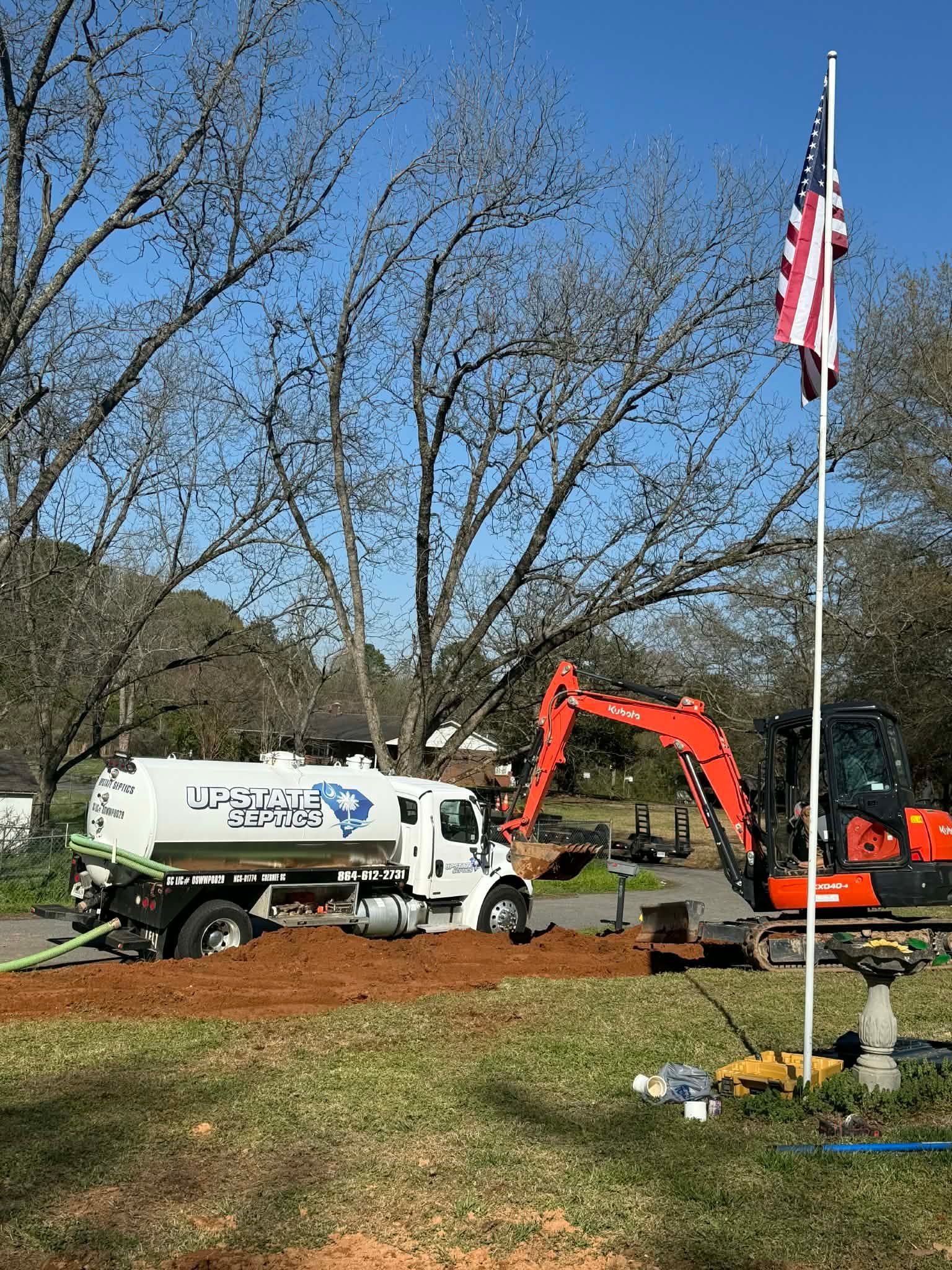 A construction scene features an orange excavator and a white water tank truck parked on a dirt lot near a US flag pole.