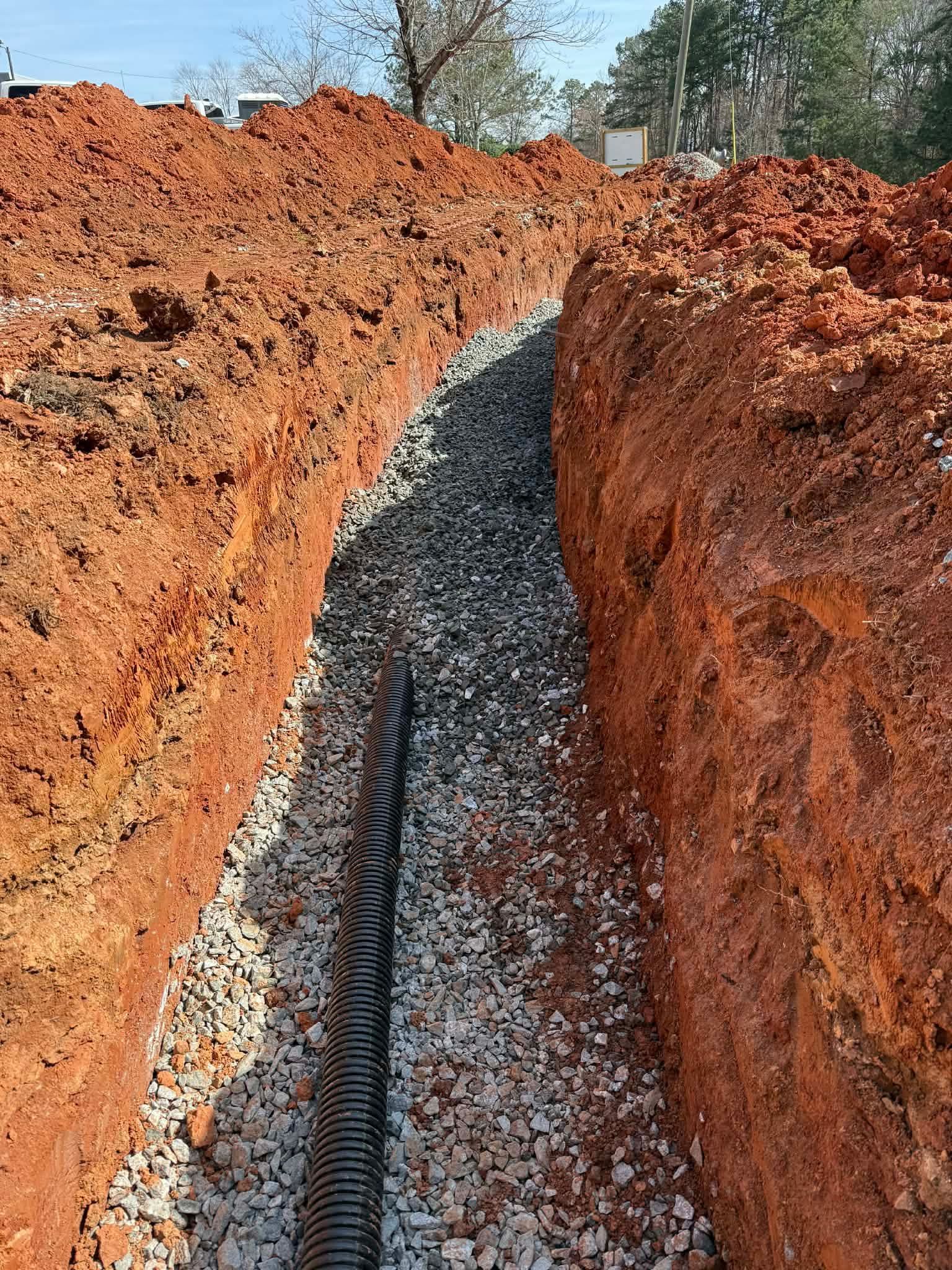 A black corrugated drainage pipe rests on a bed of gravel inside a deep, narrow trench cut into red clay soil.