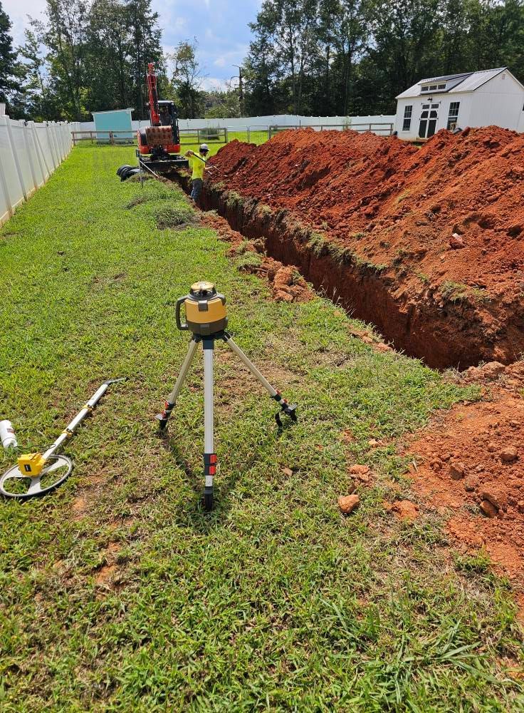 A worker uses an excavator to dig a long, deep trench in a grassy backyard, monitored by a laser level on a tripod.