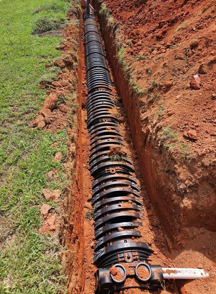 A series of black, arched plastic drainage chambers installed in a deep trench cut into reddish-brown soil.