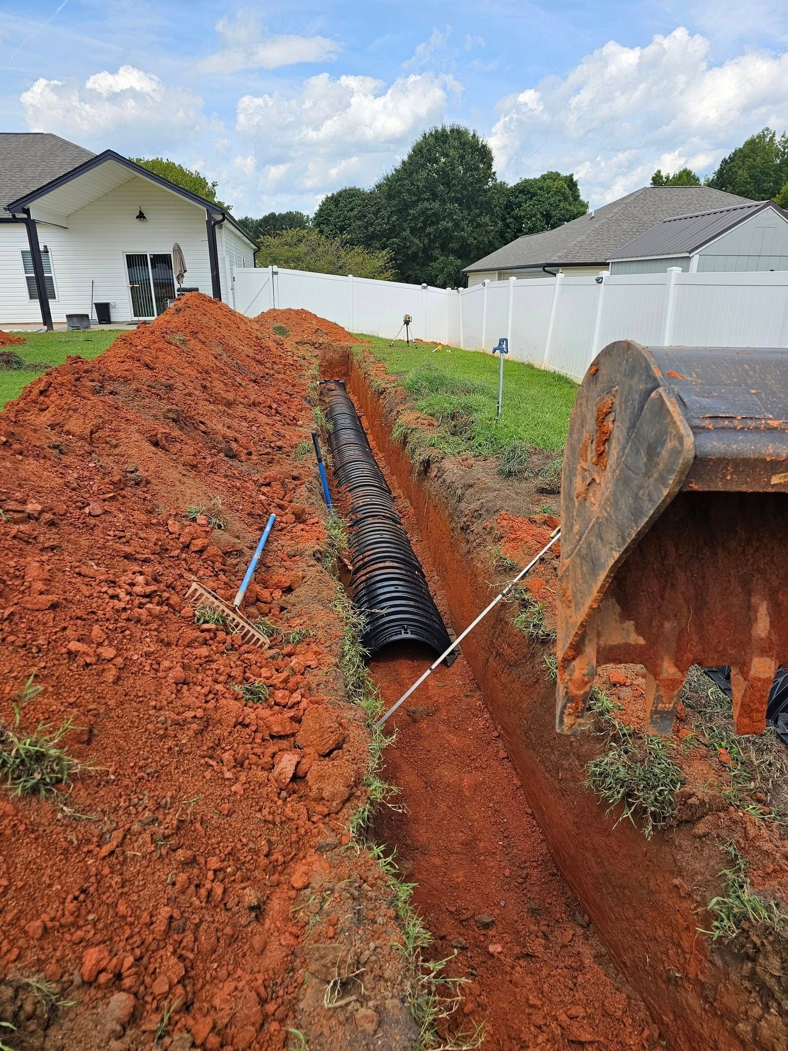 A trench containing a black drainage pipe segments in a red clay yard, with a backhoe bucket in the foreground.