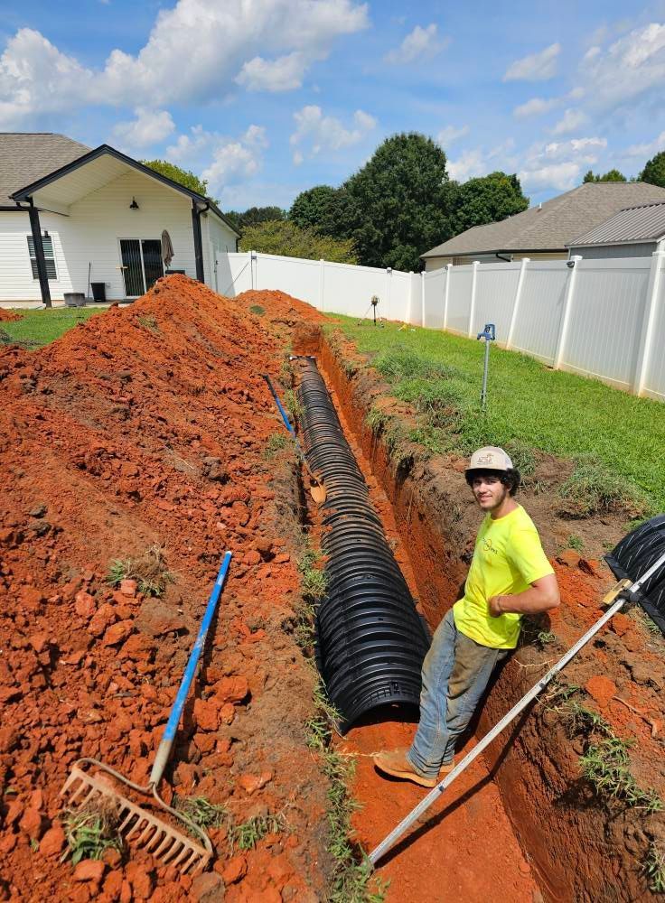 A person in a neon yellow shirt stands in a trench next to a newly installed corrugated black drainage pipe.