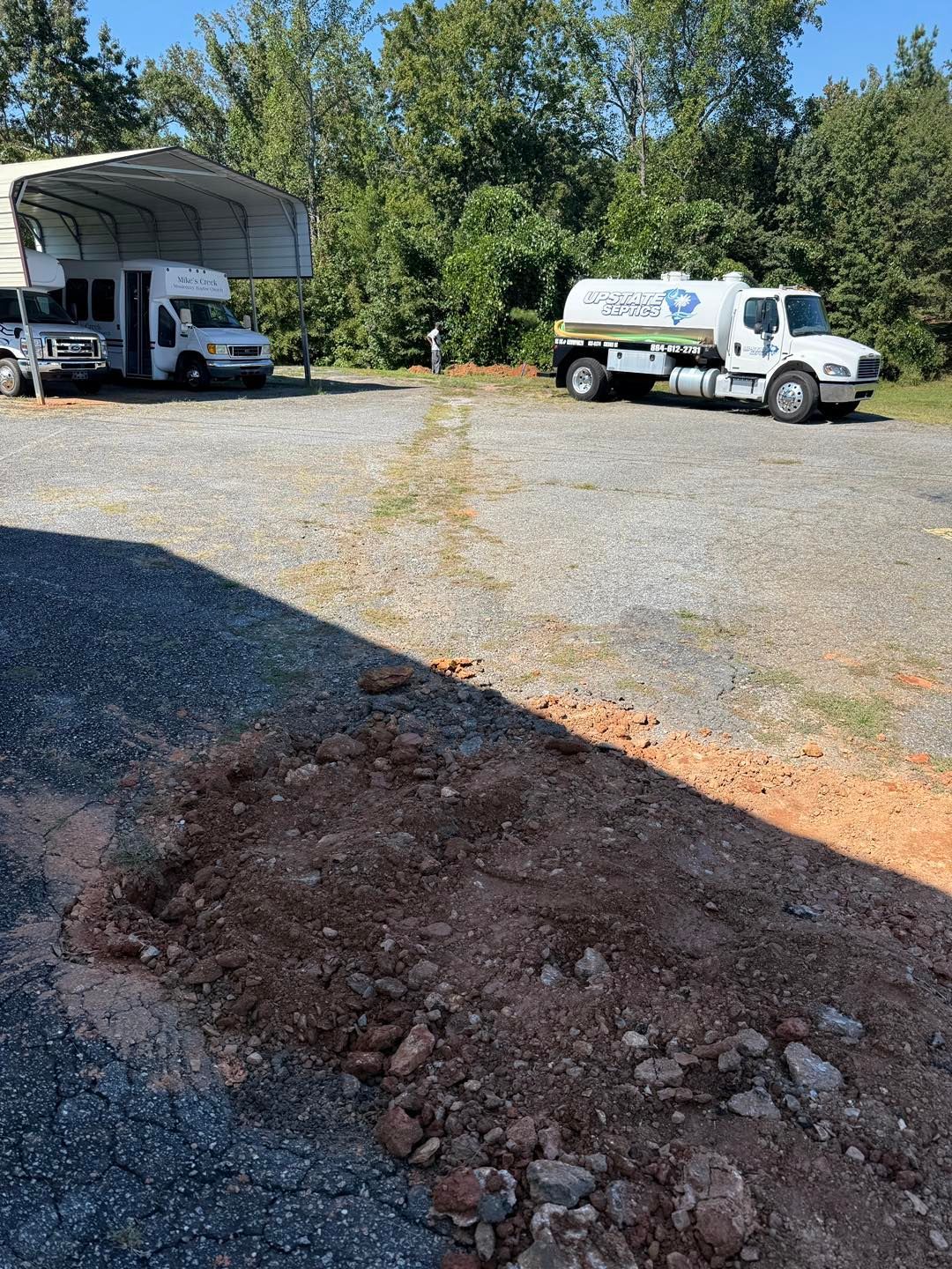 A white fuel truck parked in a gravel lot next to a covered white vehicle, with a dirt mound in the foreground.