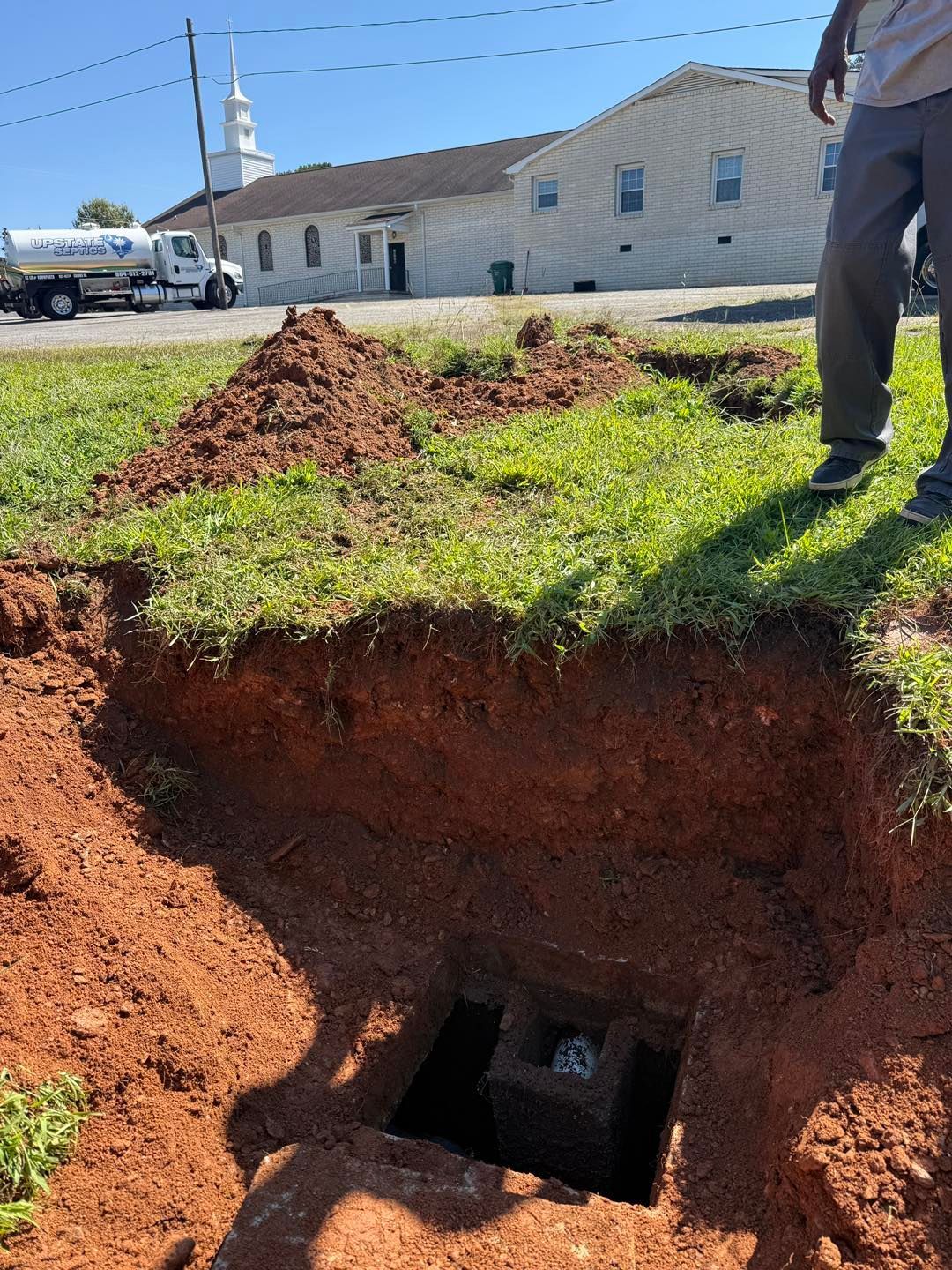 A person stands near a square pit dug into red soil, with a church and a service vehicle in the background.
