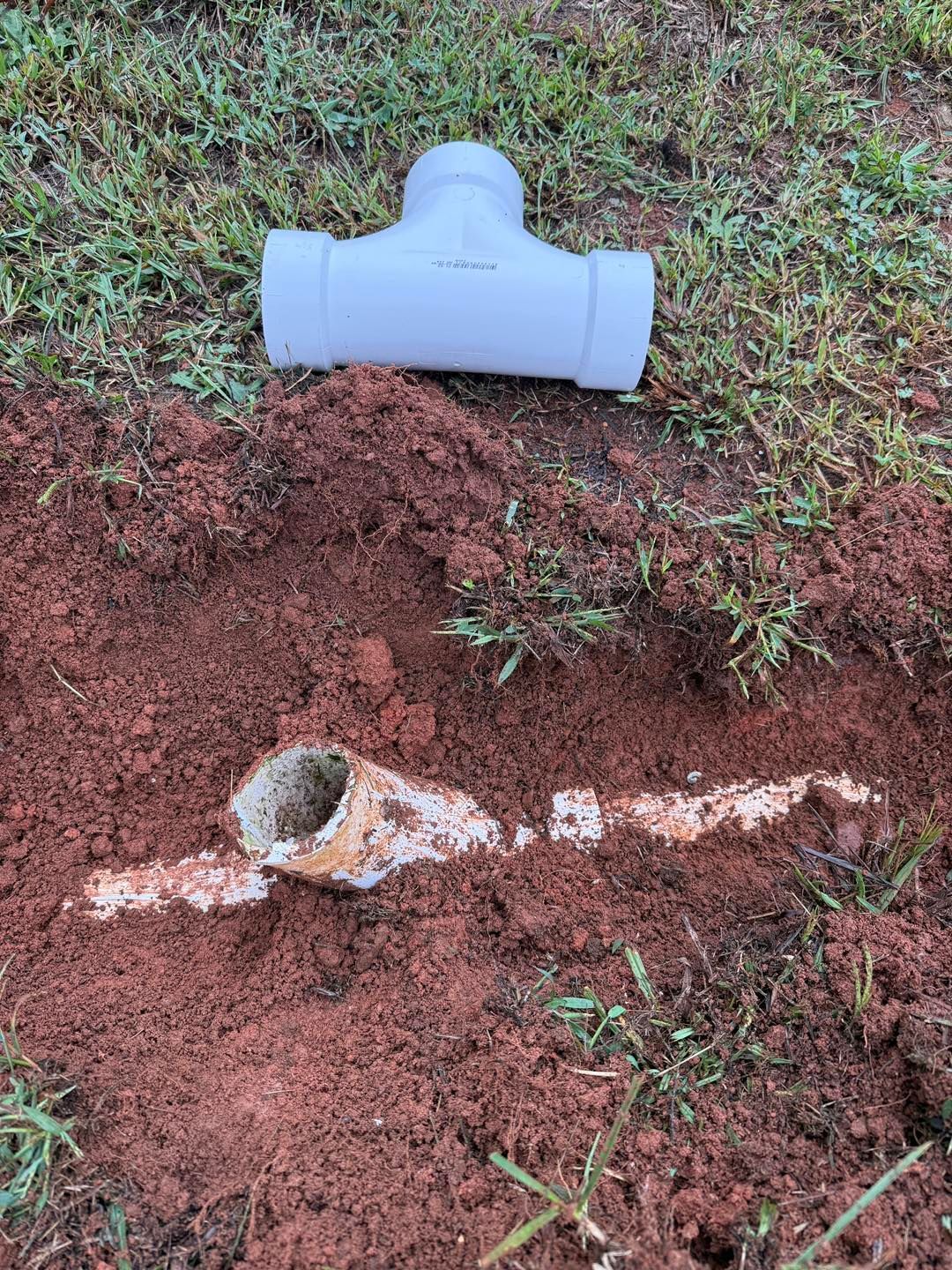A white PVC T-fitting rests on grass above a trench containing a broken, dirt-covered white pipe segment.